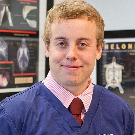 A person wearing a blue scrubs top and red tie stands smiling, in front of medical posters in a clinical or educational setting.