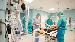 Medical staff in scrubs and surgical masks attend to a patient on an operating table in a brightly lit hospital room, surrounded by medical equipment and monitors.