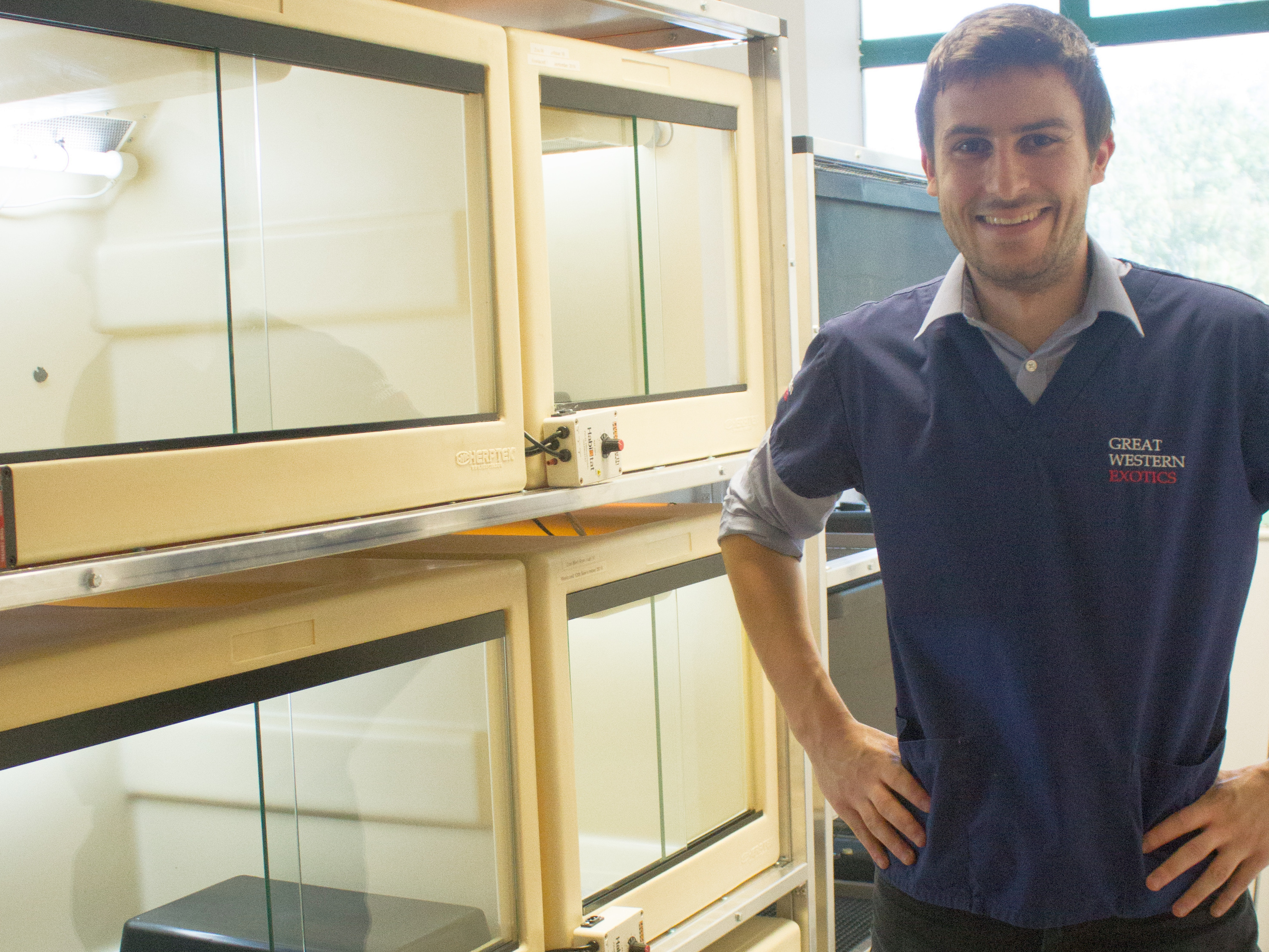 A person stands smiling beside stackable glass-fronted enclosures in a brightly lit room. They wear a shirt with GREAT WESTERN EXOTICS printed on it.