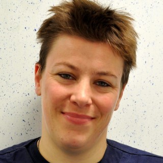Alex Tischer Image 2016 A person with short brown hair is smiling, set against a white speckled background, wearing a dark blue top.