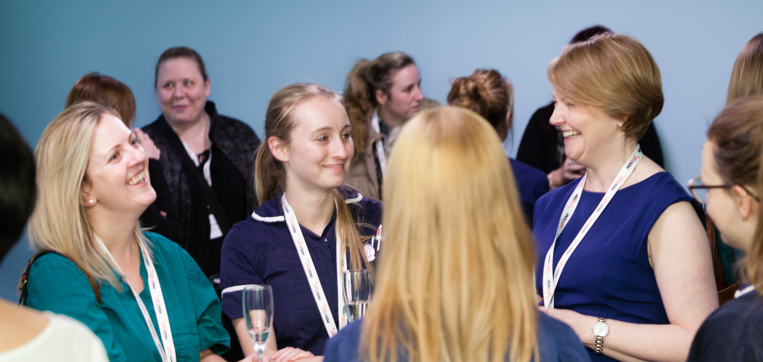 People are standing in a small group, smiling and conversing in a social gathering. They wear lanyards with RCNi printed on them, and one holds a glass, with others in the background.
