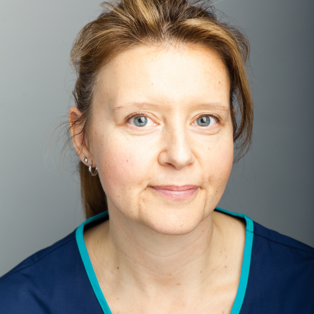 A person with light hair and blue eyes smiles subtly, wearing a navy and teal garment, set against a plain gray background.