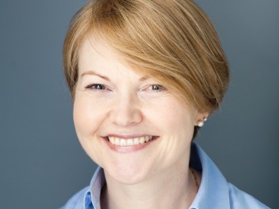 A person with short, light hair smiles warmly, wearing a light blue shirt against a plain, dark blue background.