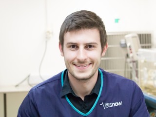 A smiling man wearing a Vets Now uniform stands indoors, possibly in a veterinary clinic, with blurred equipment in the background.