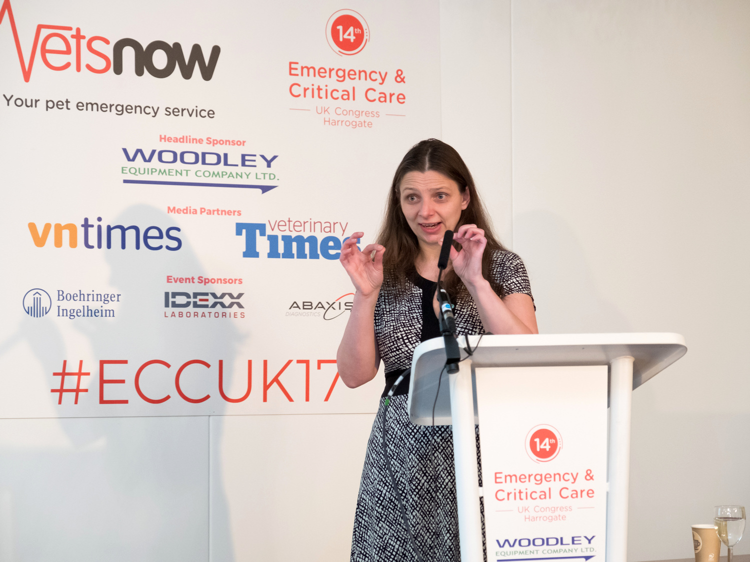Toxicology CPD A person speaks at a podium with a microphone, gesturing with their hands. The backdrop features logos and text: Vets Now, Emergency & Critical Care, ECCUK17, and sponsor names like WOODLEY EQUIPMENT COMPANY LTD.