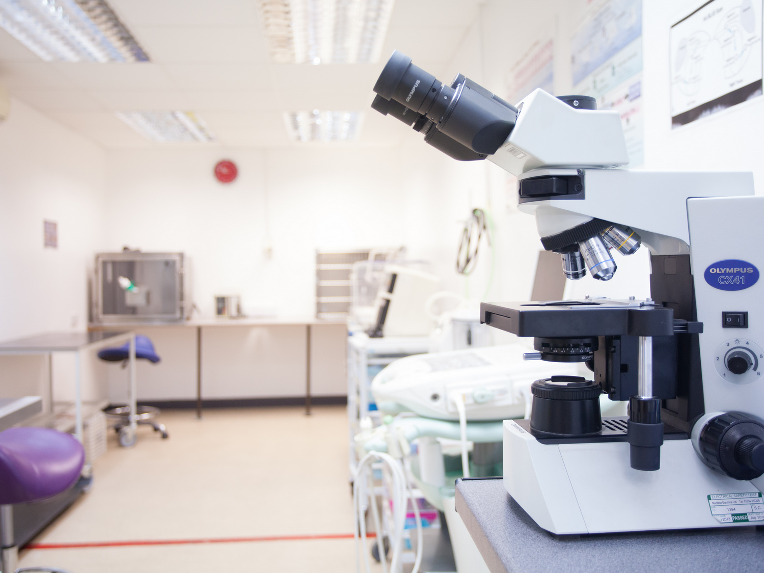 A microscope labeled Olympus CX41 is positioned on a laboratory table. The laboratory environment includes counters, a stool, and medical equipment under fluorescent lighting.