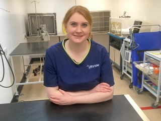 Person in blue scrubs with Vets Now logo, smiling with arms crossed, in a veterinary clinic featuring medical equipment and supplies on carts and tables.
