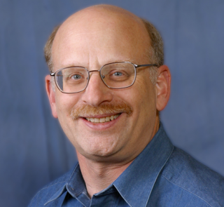A man with glasses and a mustache smiles, wearing a blue collared shirt against a plain blue background.