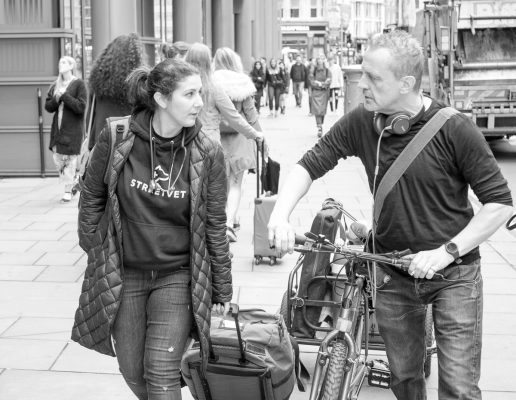 A woman and man talk while walking on a busy sidewalk. She carries bags, wearing a StreetVet hoodie. He pushes a folding bike, wearing headphones around his neck. Pedestrians fill the background.