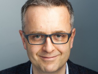 A smiling person with short hair wears glasses and a dark jacket, facing forward against a plain gray background.