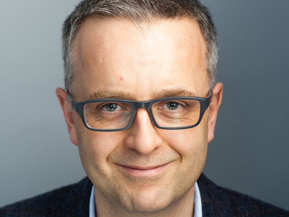 A smiling person with short hair wears glasses and a dark jacket, facing forward against a plain gray background.