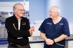 Two men are smiling and talking in a veterinary clinic. One wears a Northwest sweater, the other a John Williams FRCVS Surgery Specialist uniform. A no smoking sign is visible.