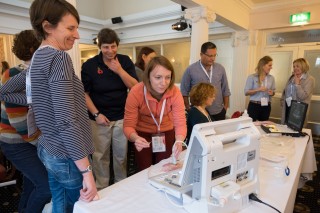 A woman in an orange sweater operates an ultrasound machine while others observe in a conference room setting with white columns and carpeted floors. Participants wear identification badges.