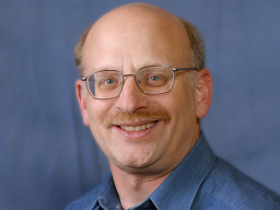 A person with glasses smiles warmly, wearing a blue shirt against a plain, blue backdrop.