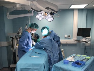 Surgeons perform an operation under bright surgical lights in a sterile, blue-painted operating room, with various medical instruments arranged on a nearby draped table.