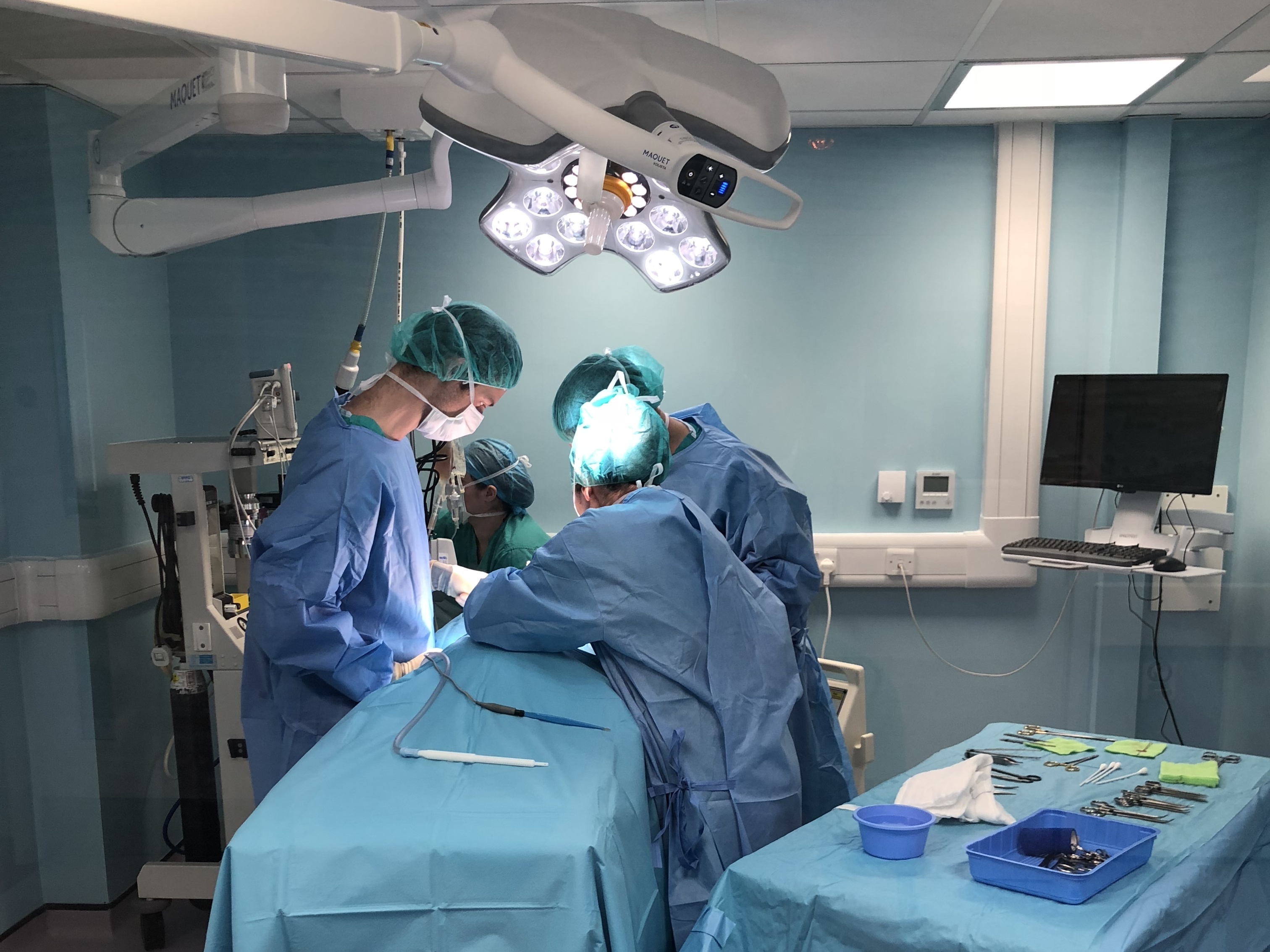Surgeons perform an operation under bright surgical lights in a sterile, blue-painted operating room, with various medical instruments arranged on a nearby draped table.