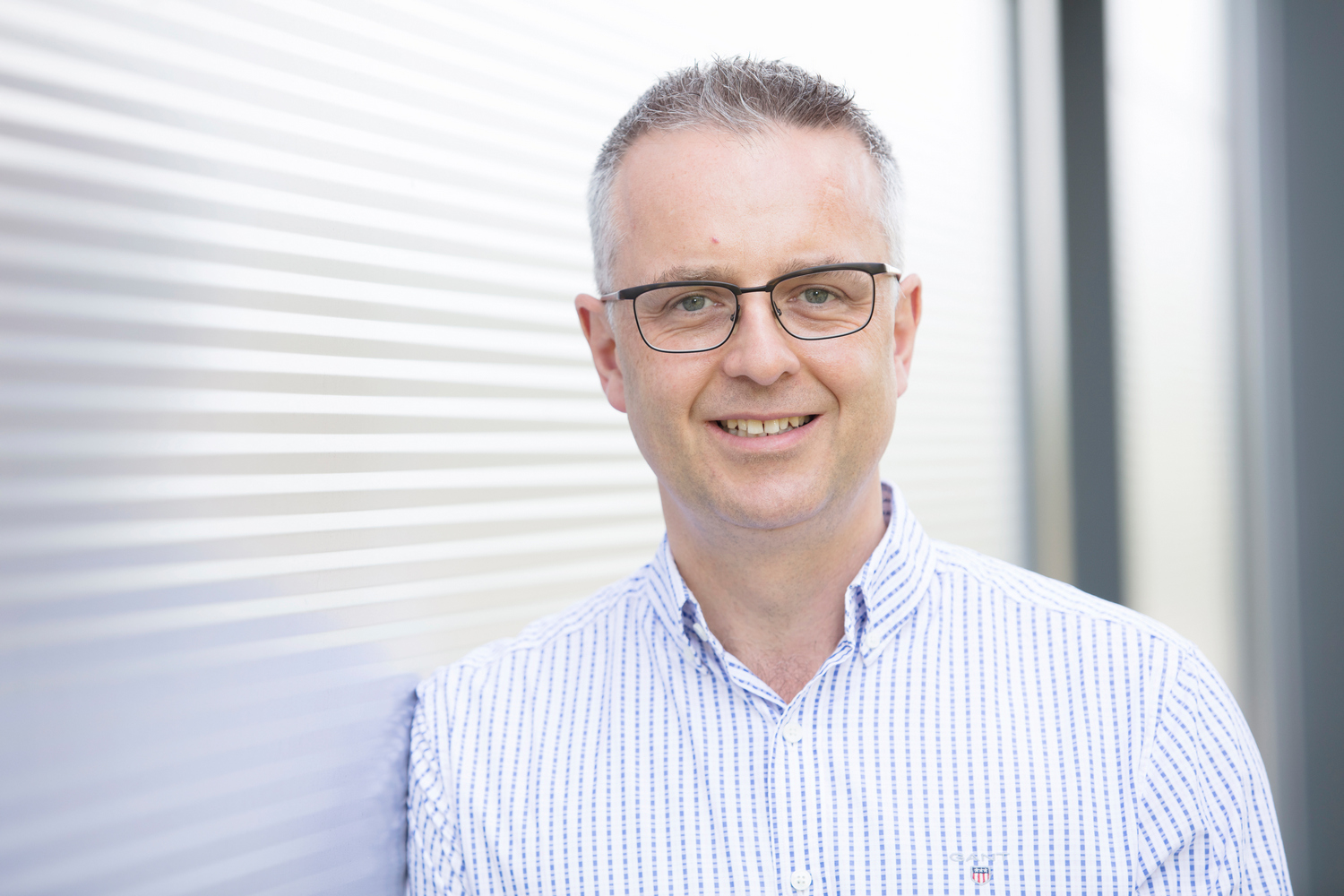 A smiling man wearing glasses and a striped shirt stands against a corrugated metal wall, creating a casual, professional setting.