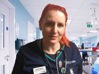 A nurse with red hair, wearing a stethoscope and a name tag reading Lesley Moore Principal Nurse Manager Vets Now, stands in a clinical setting with monitors and medical equipment.