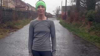 A person stands on a wet, rural road wearing a gray outfit and green headband. Houses and leafless trees line the street under an overcast sky.