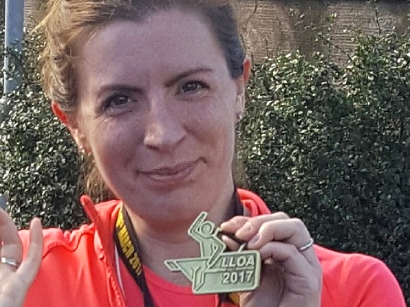 A person holds a medal with ALLOA 2017 engraved, standing outdoors in front of greenery, wearing a bright jacket and a lanyard around the neck.