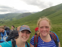 Two smiling women with backpacks stand in a group on a grassy hillside under a cloudy sky, with rolling green hills in the background. Cap reads ARIAT SINCE 1993.