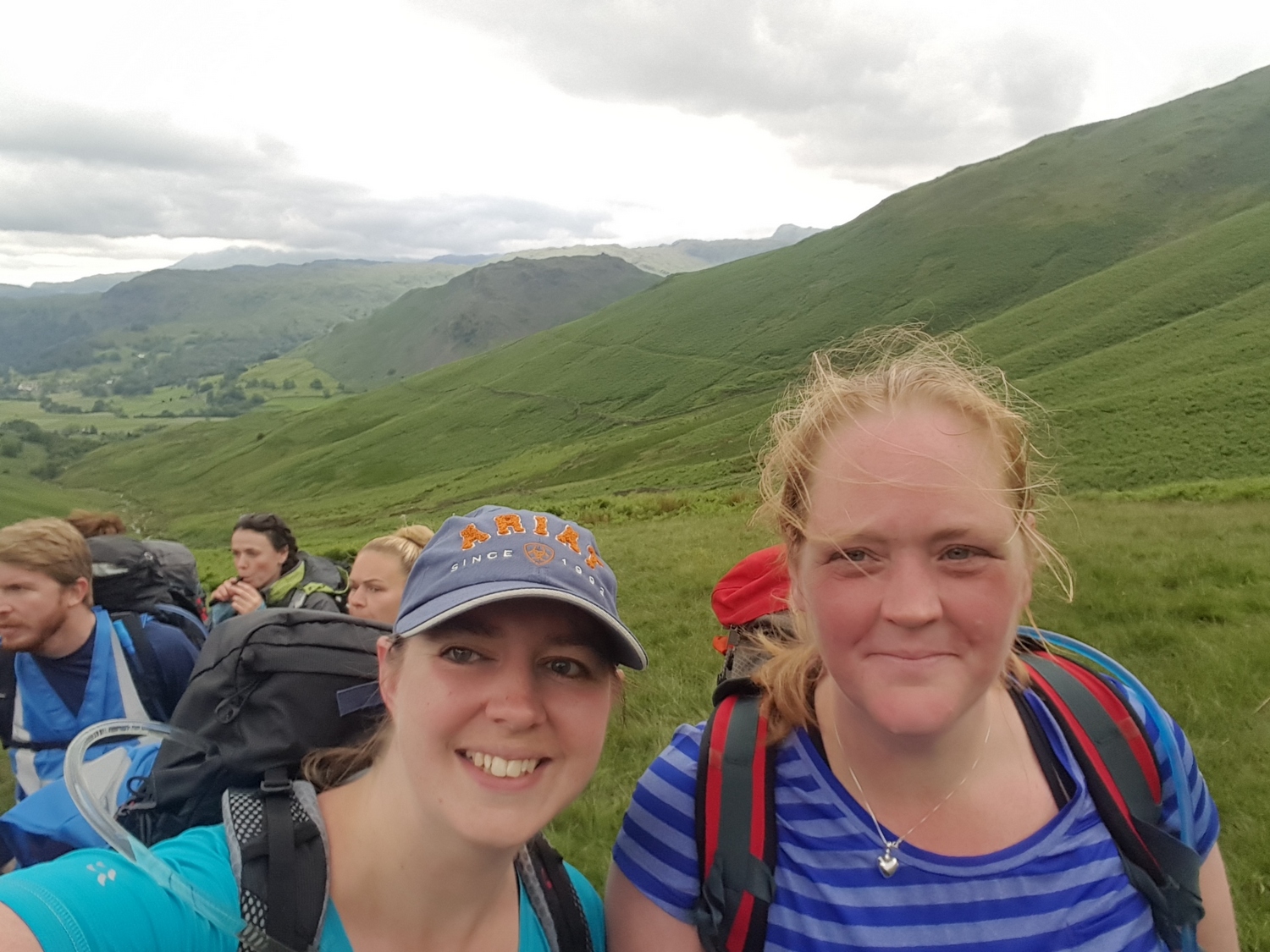 Two smiling women with backpacks stand in a group on a grassy hillside under a cloudy sky, with rolling green hills in the background. Cap reads ARIAT SINCE 1993.