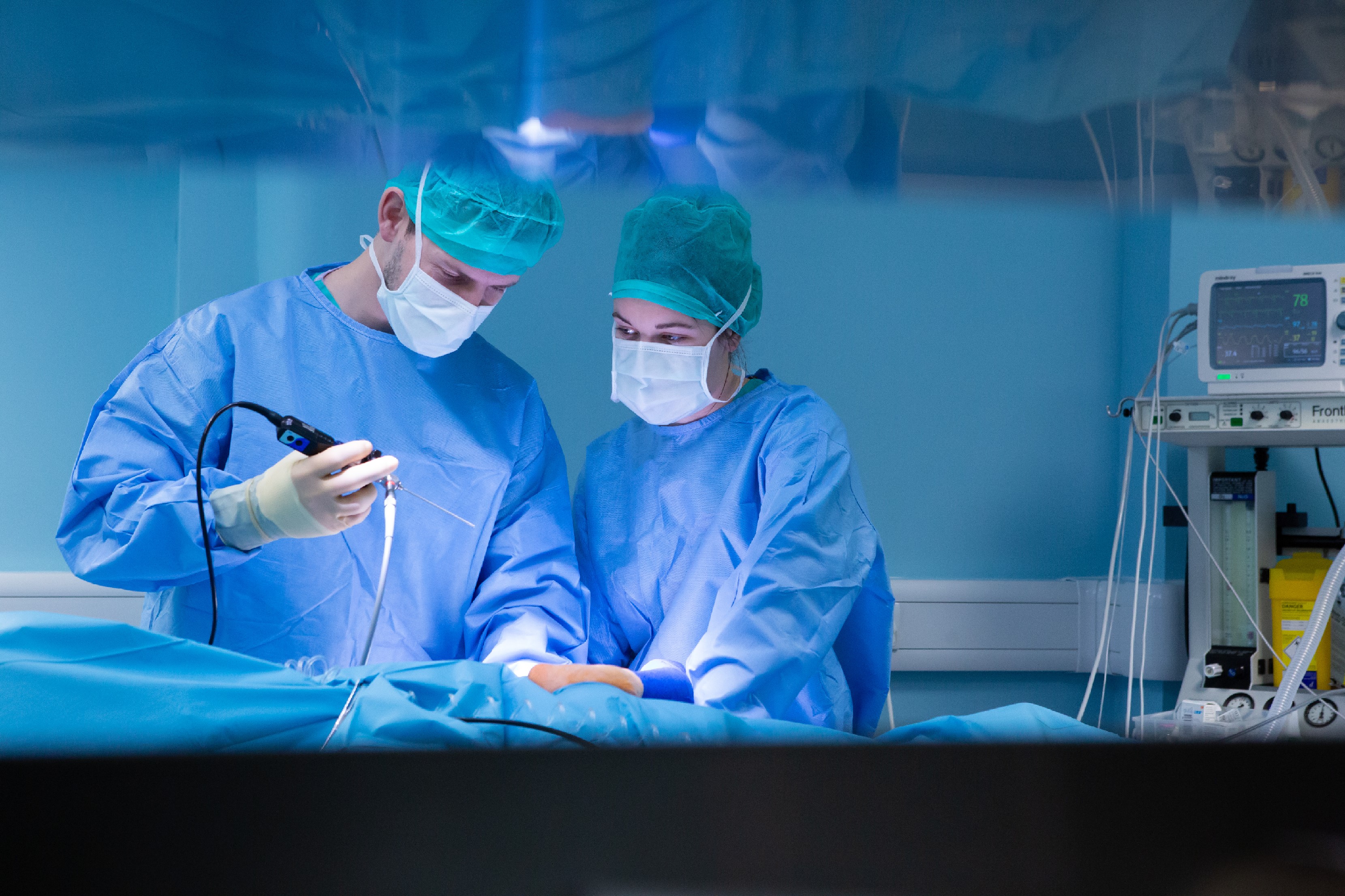 Two surgeons wearing blue scrubs and masks use a surgical instrument while operating in an operating room, surrounded by medical equipment and monitors.