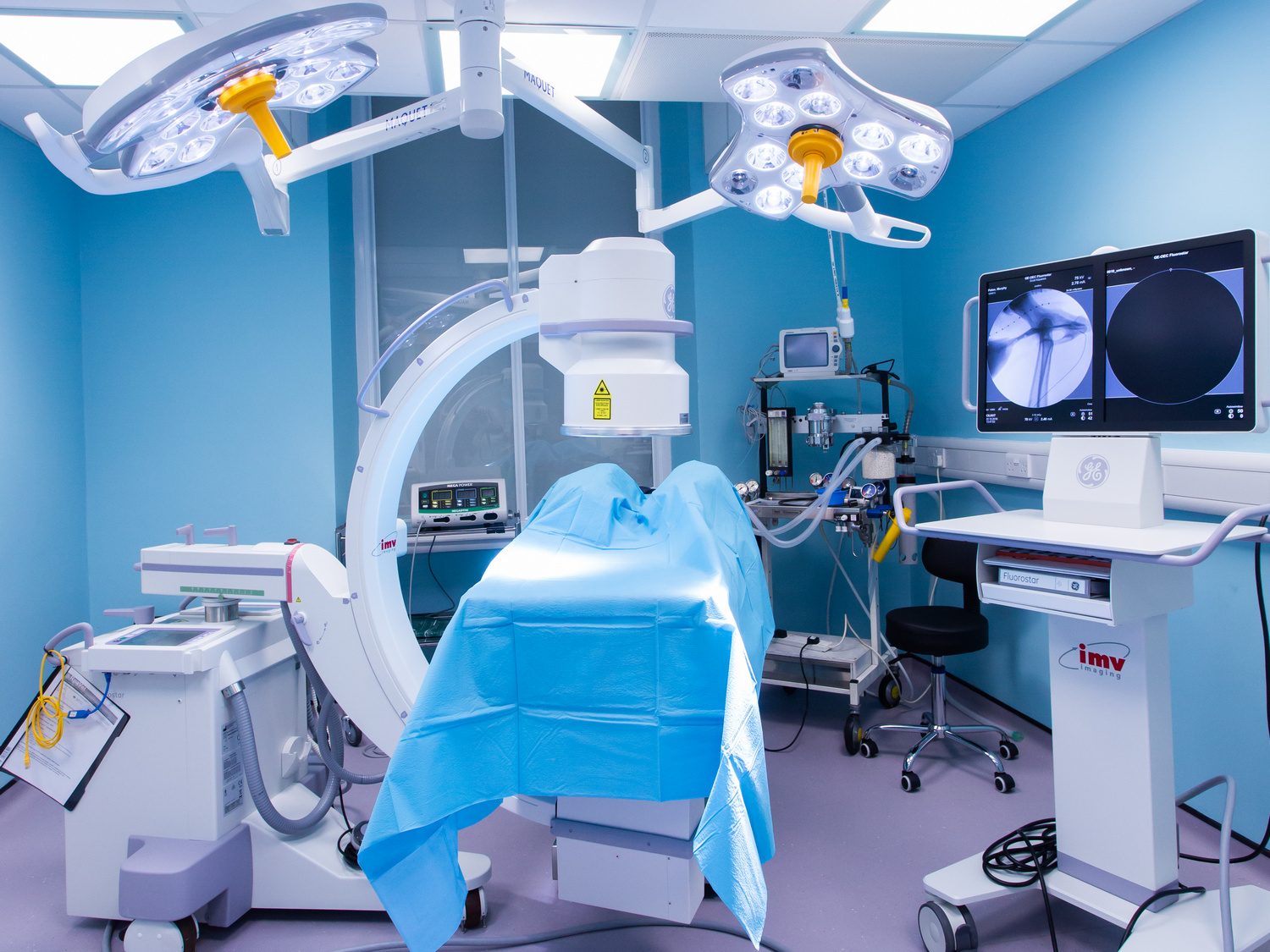 A hospital operating room features a C-arm X-ray machine positioned over a surgical table, surrounded by high-tech equipment, monitors displaying medical images, and overhead LED surgical lights.