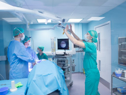 Medical staff work around an operating table under bright lights; the lead surgeon adjusts equipment while others observe, within a sterile, blue-lit surgical room.
