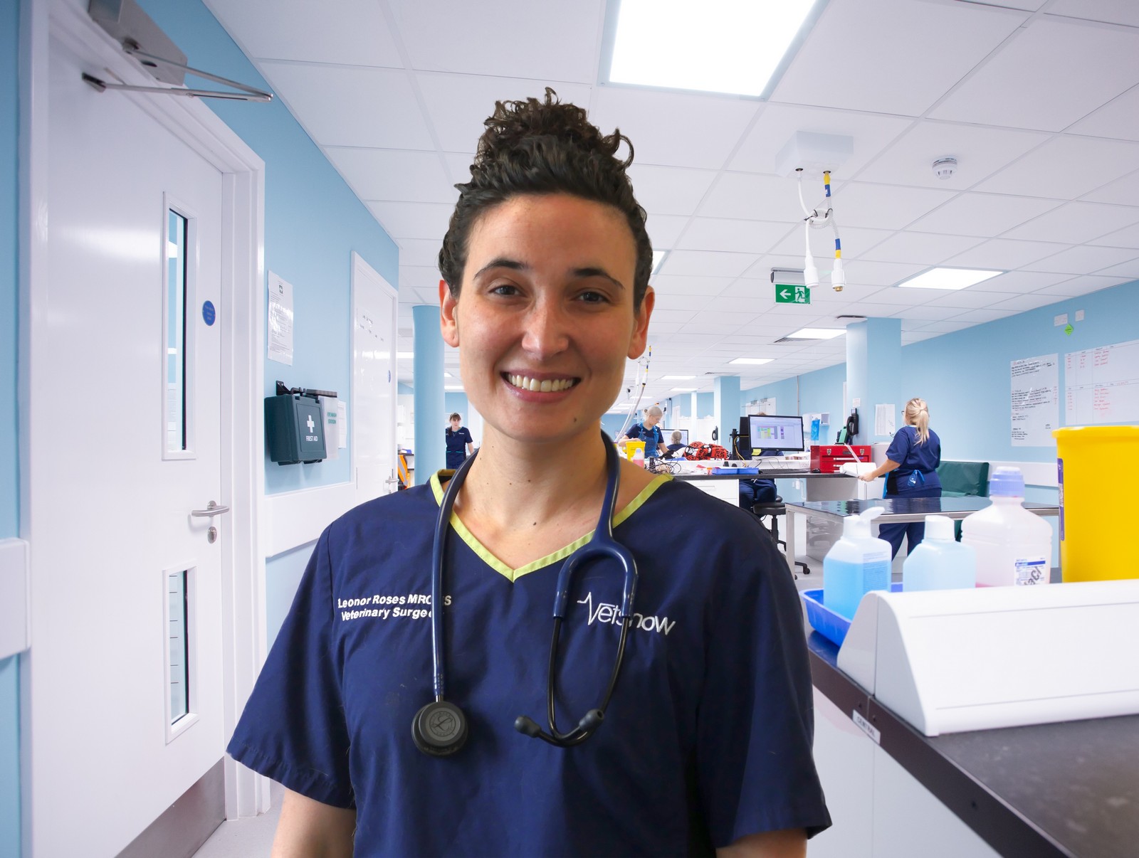 A person wearing scrubs with a stethoscope smiles in a bright, blue veterinary clinic. The text reads: Leonor Rosas MRCVS, Veterinary Surgeon and Vetsnow on the uniform.