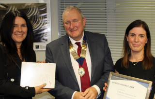 Three people stand smiling indoors; the man wears a medallion and suit, flanked by two women holding a box and an award certificate. Text: DESIGN AWARDS 2019 presented to Vets Now Emergency Ltd, Winner in the Refurbishment Category.