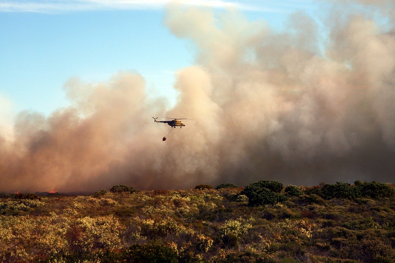 A helicopter carries a water bucket, flying over dense smoke from a wildfire engulfing a dry, bushy landscape during daylight.