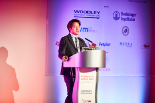 A speaker delivers a presentation at a podium labeled Emergency & Critical Care. Logos for sponsors like Woodley Equipment, Boehringer Ingelheim, and Petplan are visible in the conference setting.