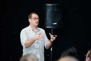 A person wearing glasses gestures while speaking, holding a remote, against a black background with a speaker. The name tag reads, “Daniel Lewis, Web Now.”