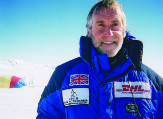 Miles_Hilton_Barber A man in a blue expedition jacket smiles in a snowy landscape. The jacket displays patches: a UK flag, RNIB Miles Hilton-Barber South Pole Expedition 2000-2001, DHL Worldwide Express, and PHD Mountain Software.