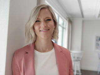 A person with short blonde hair smiles while wearing a pink blazer and white shirt, standing in a bright room with large windows and white furniture in the background.