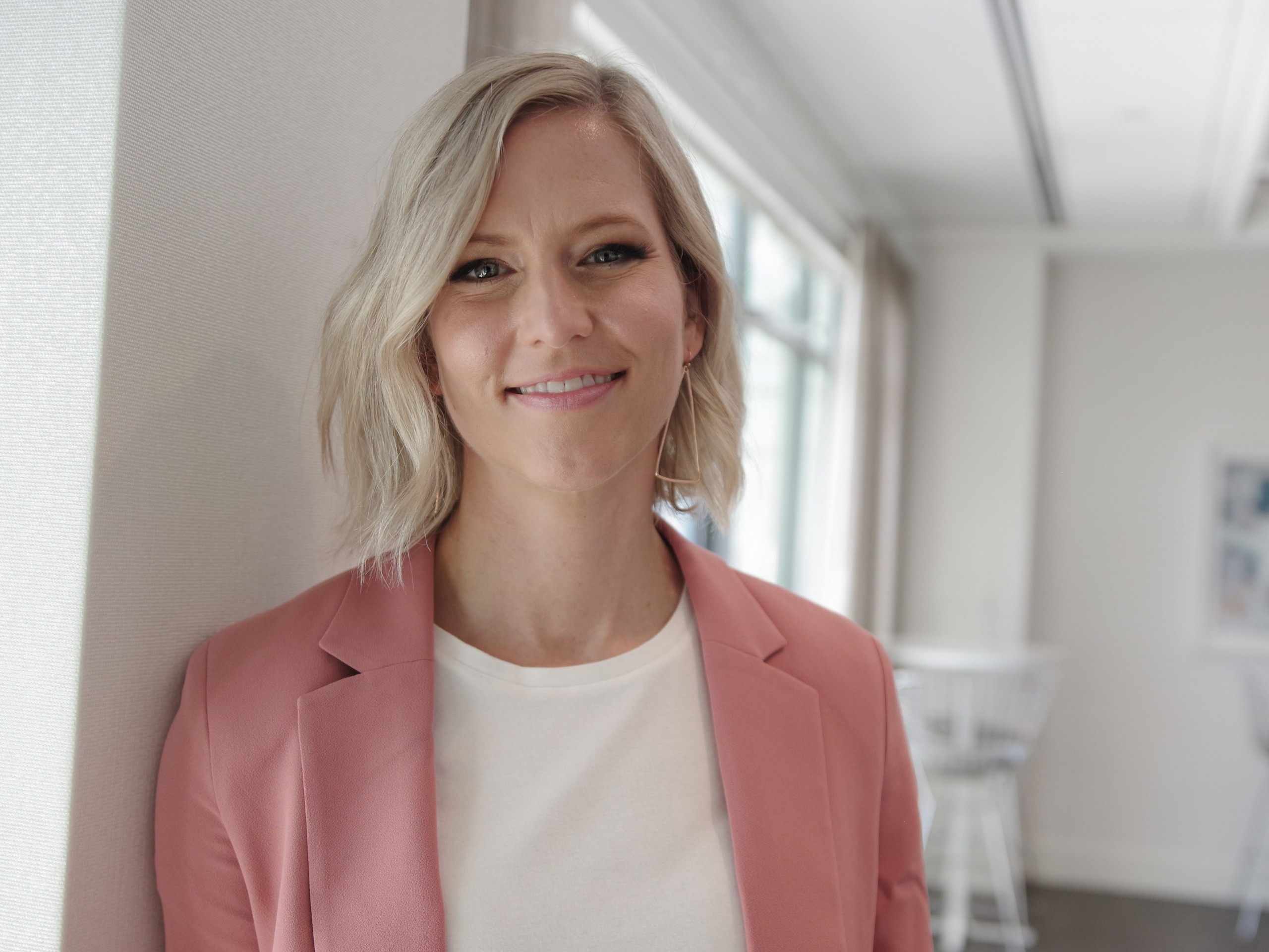 A person with short blonde hair smiles while wearing a pink blazer and white shirt, standing in a bright room with large windows and white furniture in the background.