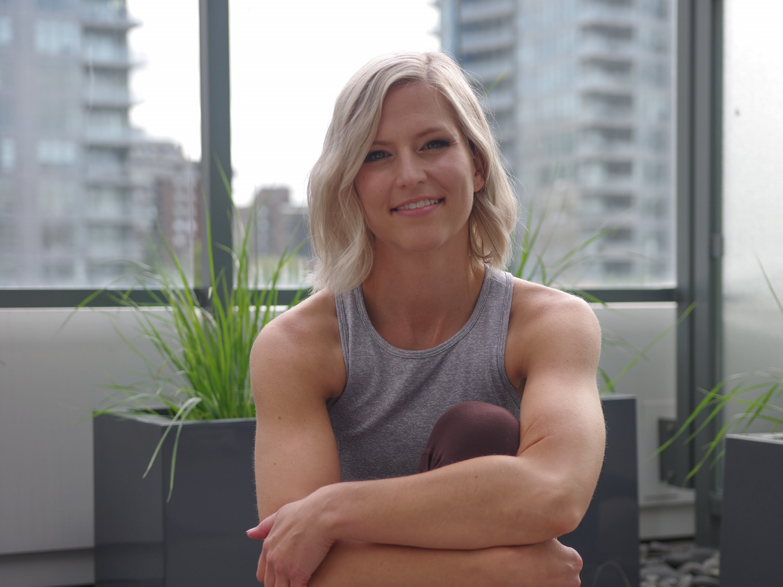 A person with light hair smiles while sitting on a balcony, surrounded by potted plants and urban high-rise buildings visible in the background.