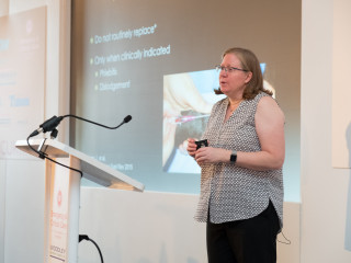A person presents in front of a projected slide. Text on slide: Do not routinely replace* Only when clinically indicated Phlebitis Dislodgement. A microphone stands nearby on a podium.