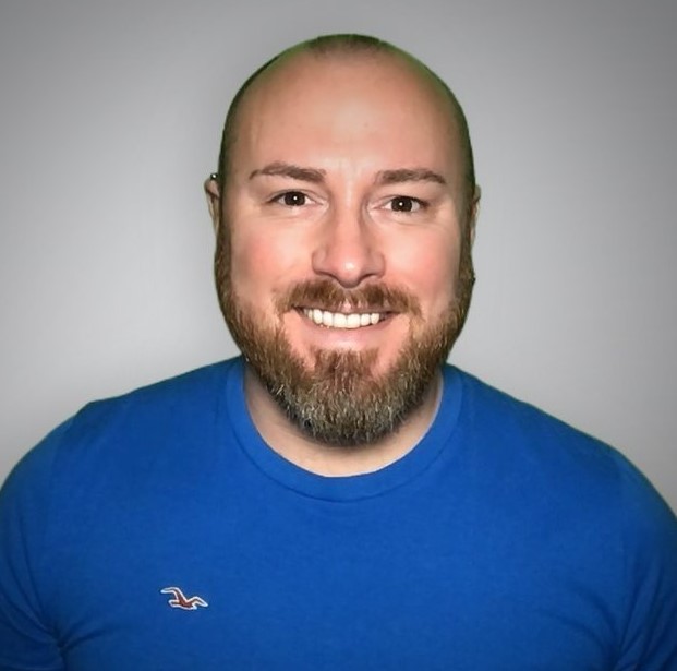 A bearded man smiles while wearing a blue T-shirt with a small bird logo, set against a plain gray background.