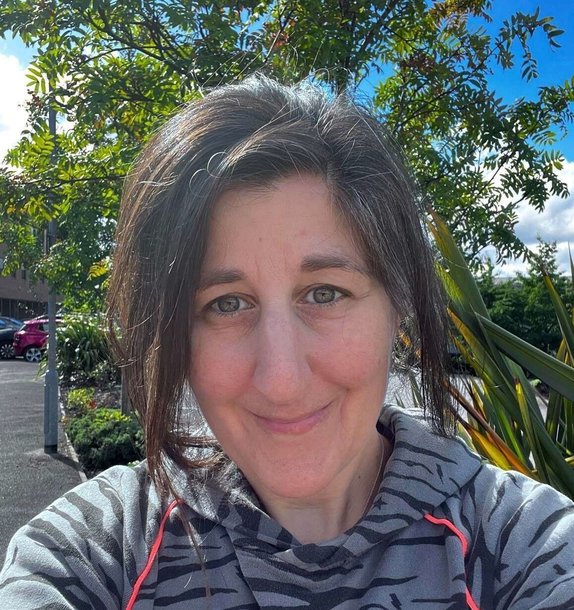 A person with dark hair stands smiling outdoors, wearing a zebra-patterned hoodie. Behind them, a tree with green leaves is visible under a clear, sunny sky.