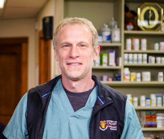 A person wearing medical scrubs and a vest stands smiling in a veterinary clinic, with shelves of various medications and supplies in the background. The vest shows University of Calgary Veterinary Medicine.