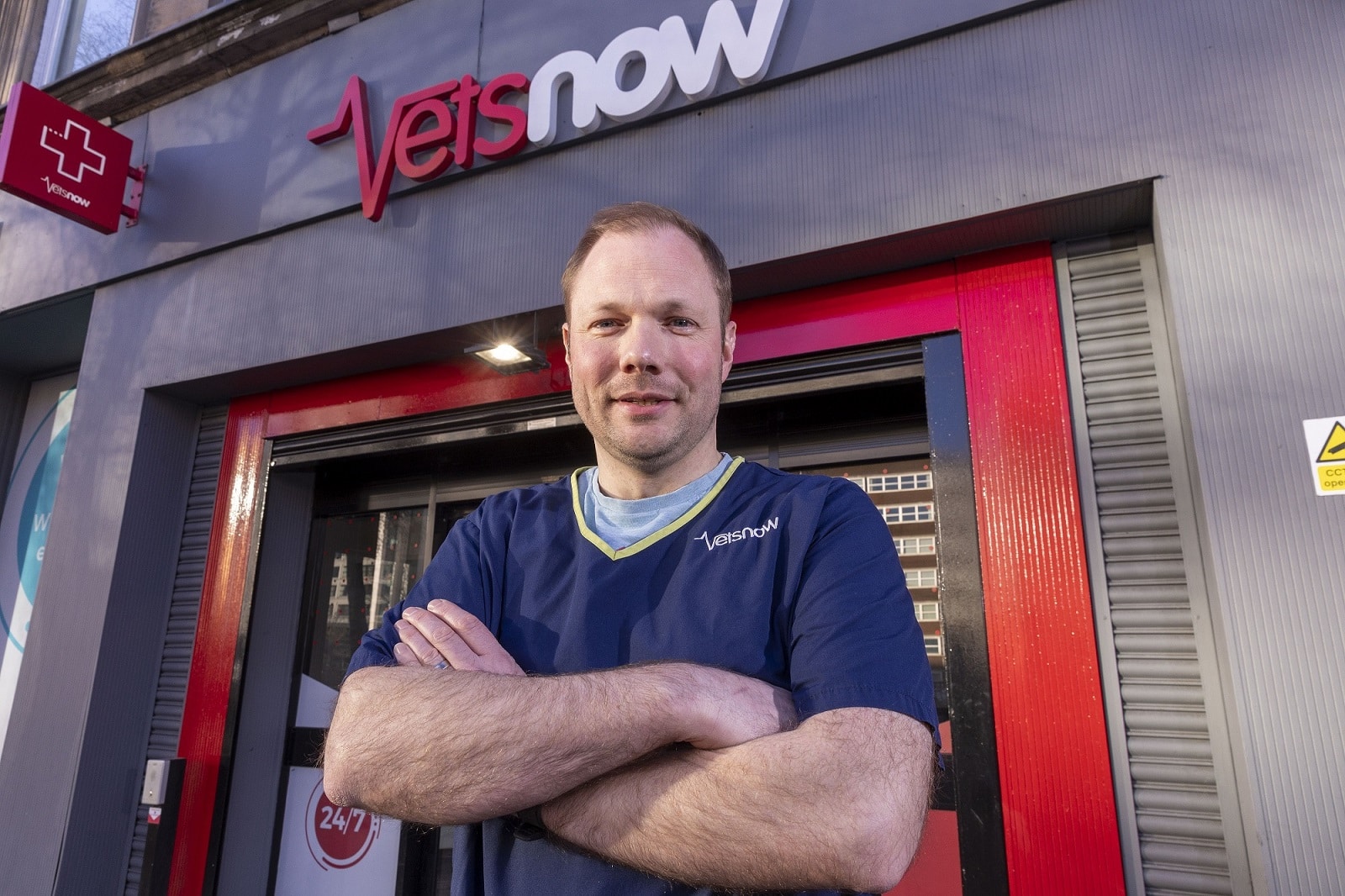 A person stands confidently with crossed arms, wearing a blue Vets Now uniform, outside a veterinary clinic with visible signs: Vets now and a red cross logo.