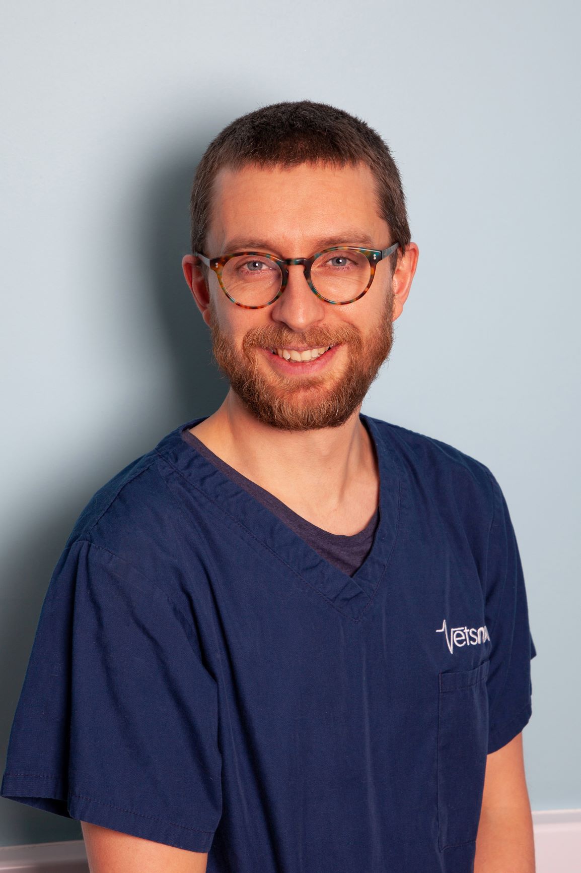 Chris A person with glasses is smiling, wearing blue scrubs with Vets written on them, against a light blue wall backdrop.