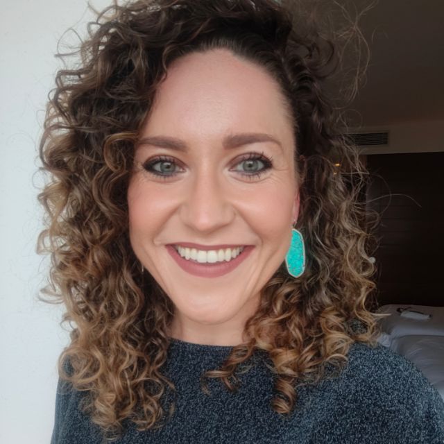 A woman with curly hair smiles at the camera, wearing a turquoise earring, in a warmly lit indoor setting with a white wall and dark furnishings in the background.