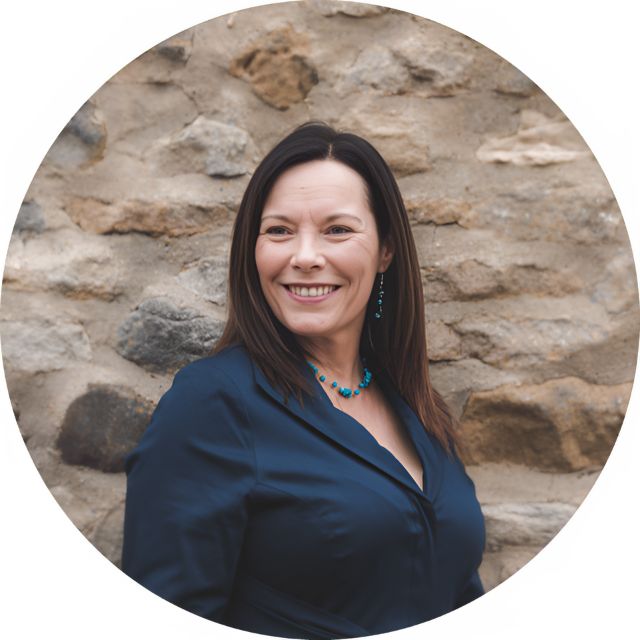 A woman in a dark blue blouse is smiling while standing against a stone wall, wearing turquoise earrings and a necklace.