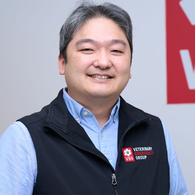 A person smiles while wearing a black vest with VEG Veterinary Emergency Group logo. They stand indoors against a white wall featuring partial red signage.