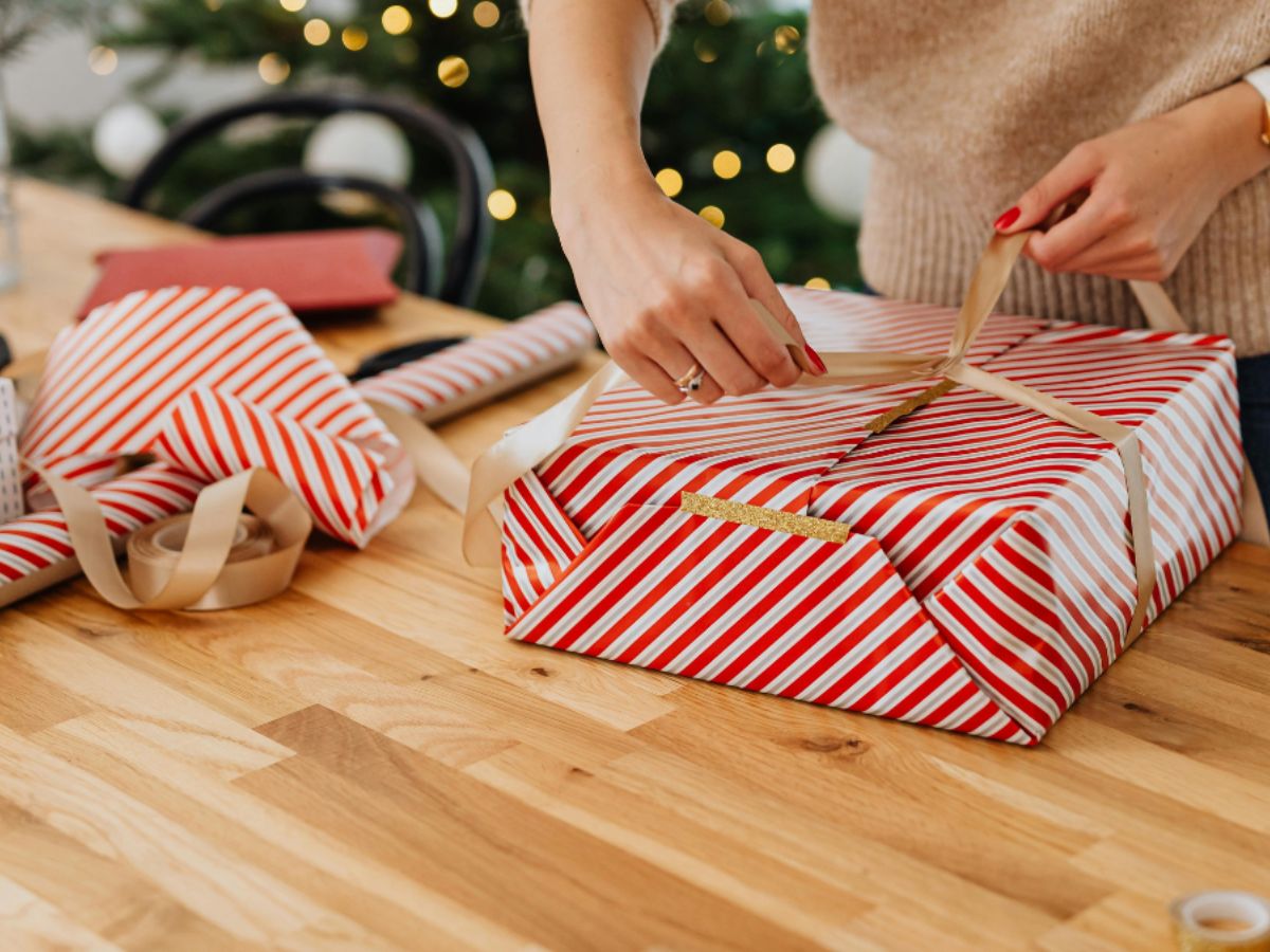 christmas-present-wrapping-hands Woman wrapping a Christmas gift in red and white striped paper with gold ribbon on wooden table