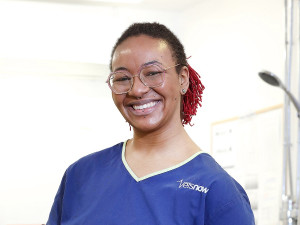 A Vets Now Vet wearing blue scrubs smiles against a plain white background.