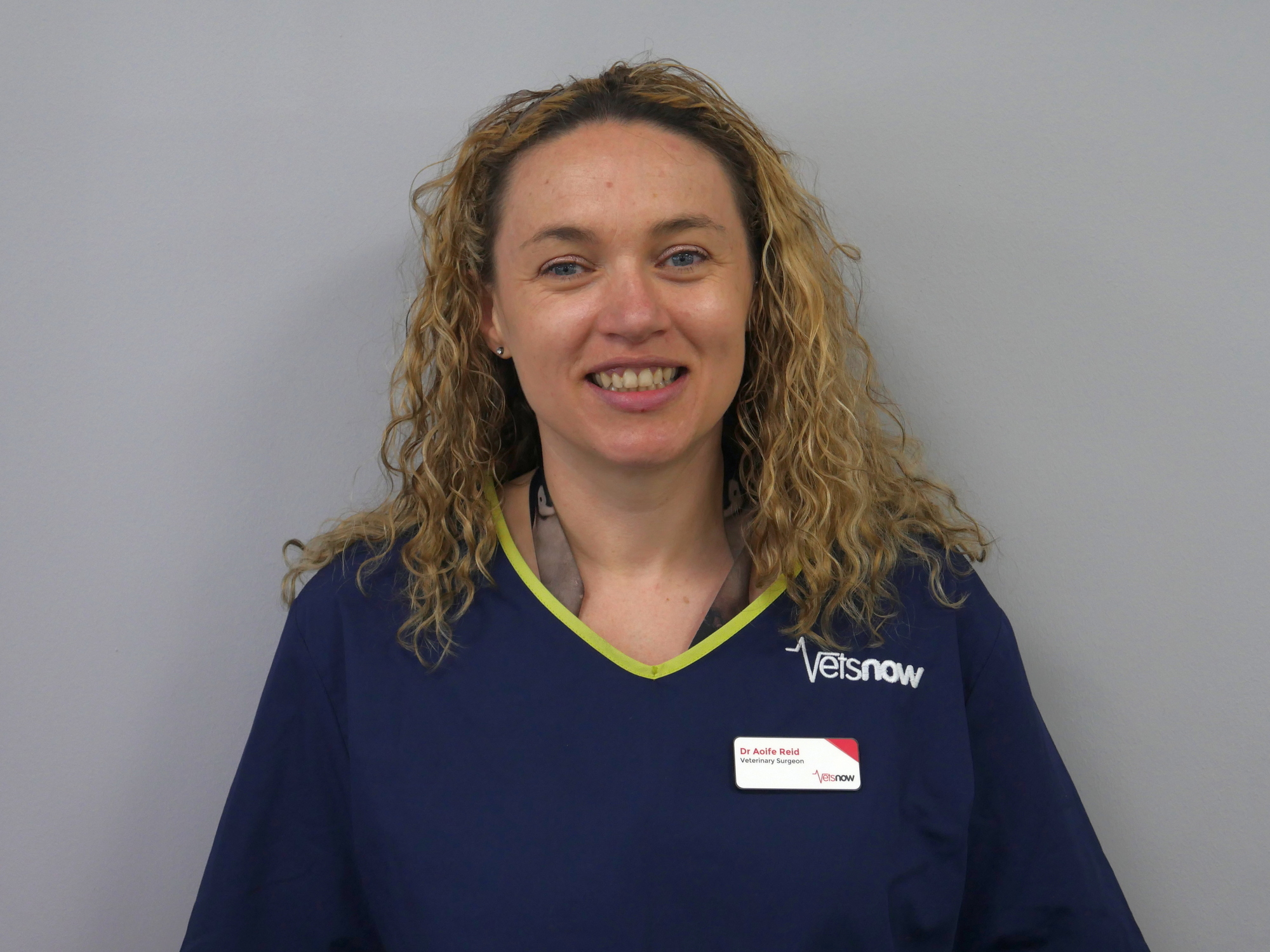 A woman with curly hair smiles, wearing a navy blue uniform with “Vetsnow” logo and a name tag reading “Dr Aoife Reid, Veterinary Surgeon,” against a plain gray background.